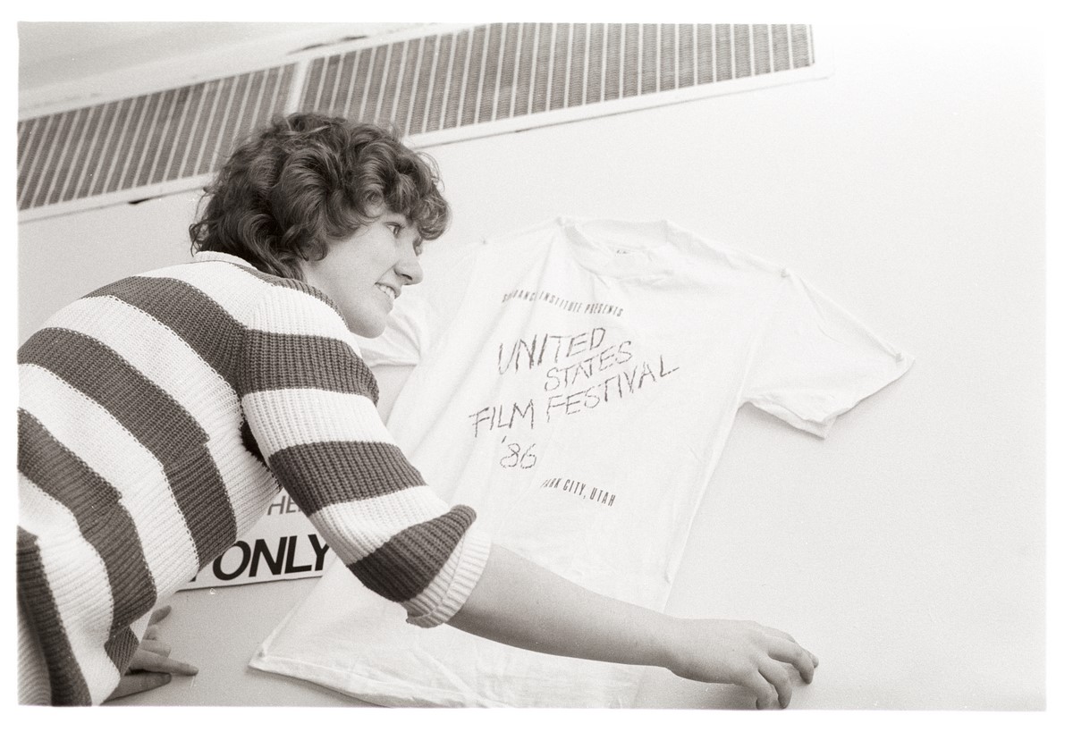 A black and white photo of a short-haired woman with a striped sweater hanging up a Festival tee that reads: United States Film Festival 86'