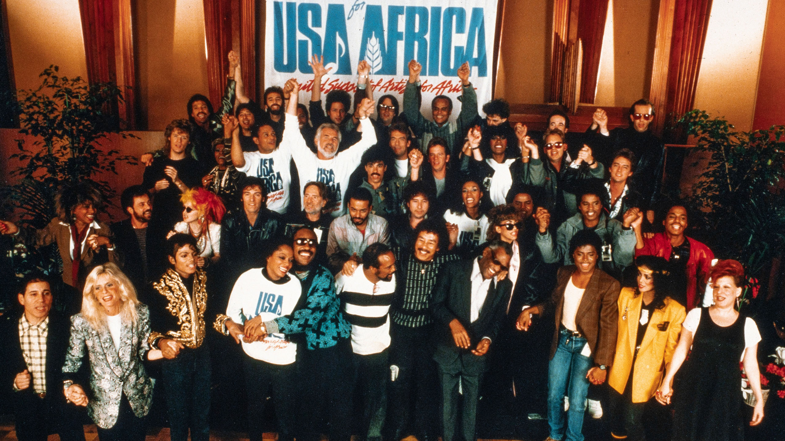A film still from The Greatest Night in Pop that shows a large group of artists assembled under a sign for USA for Africa.